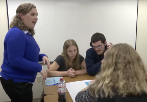 A female teacher standing with a group of students around a table in a classroom.