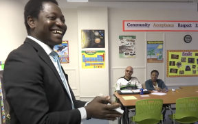 A male teacher standing before students sitting at a table in a classroom.